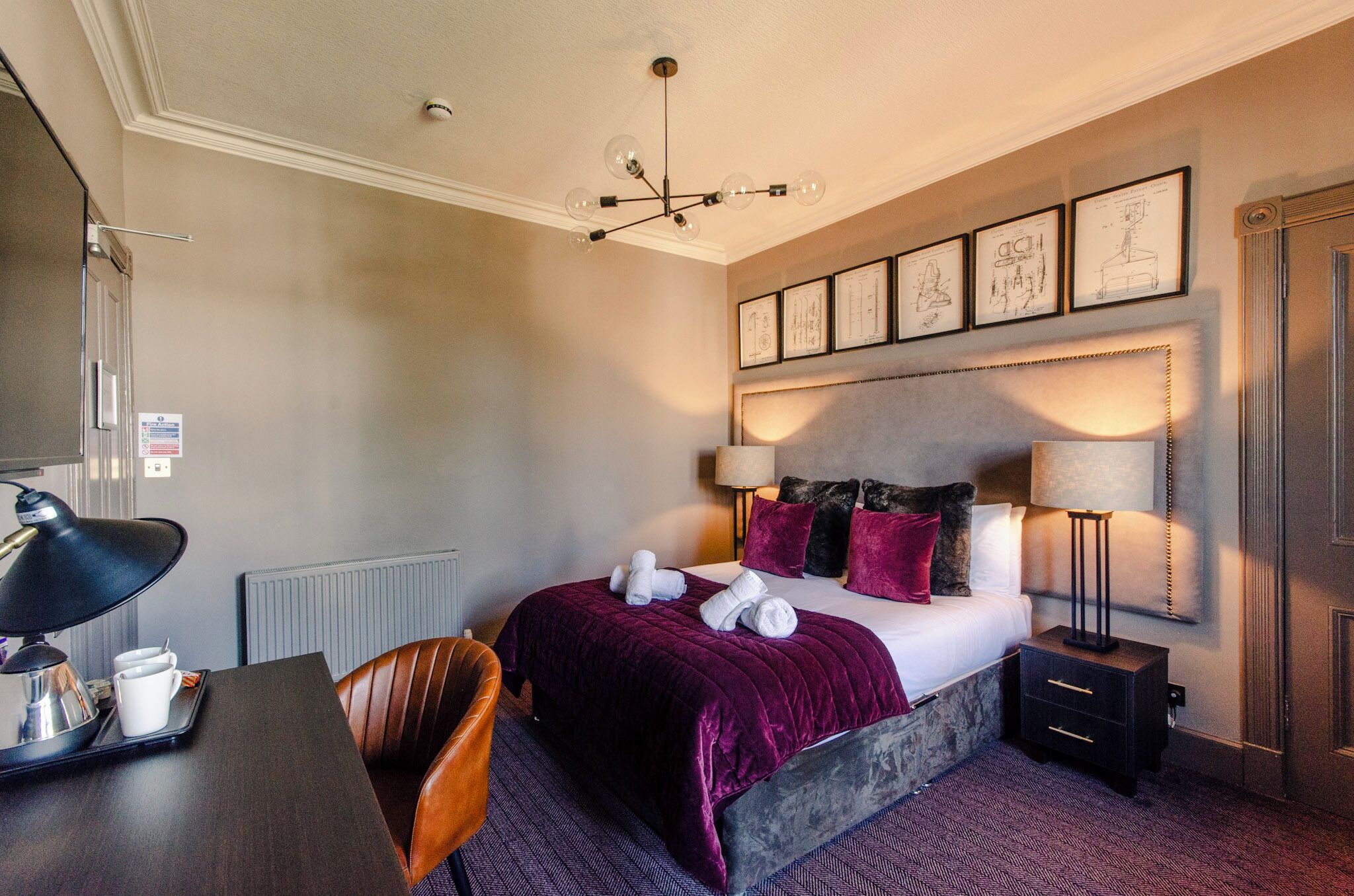 A hotel room with a bed, bedside lamps, a desk with a lamp, a chair, and framed artwork on the wall.