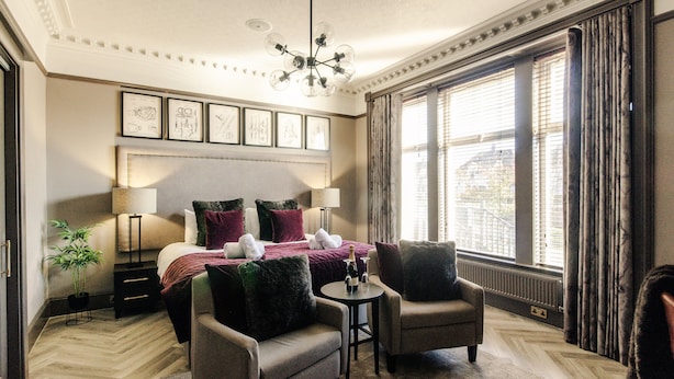 Une chambre d’hôtel avec un grand lit, deux fauteuils, une petite table, un lustre et de grandes fenêtres avec des rideaux.