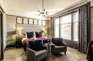 A hotel room with a large bed, two armchairs, a small table, a chandelier, and large windows with curtains. - Sweeney Aviemore Two (Aviemore)