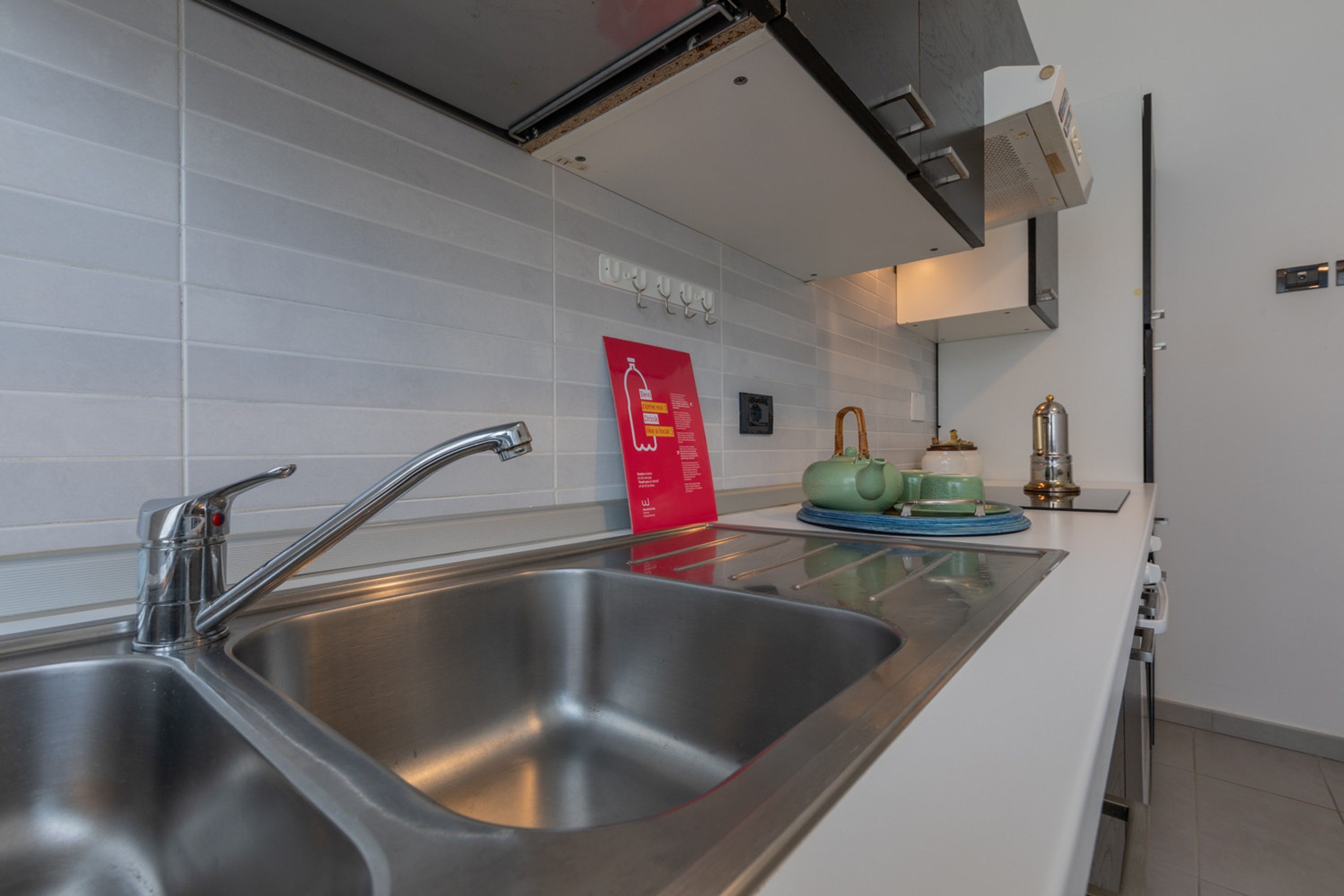 A modern kitchen with stainless steel sinks, a white tiled backsplash, and a red informational poster.
