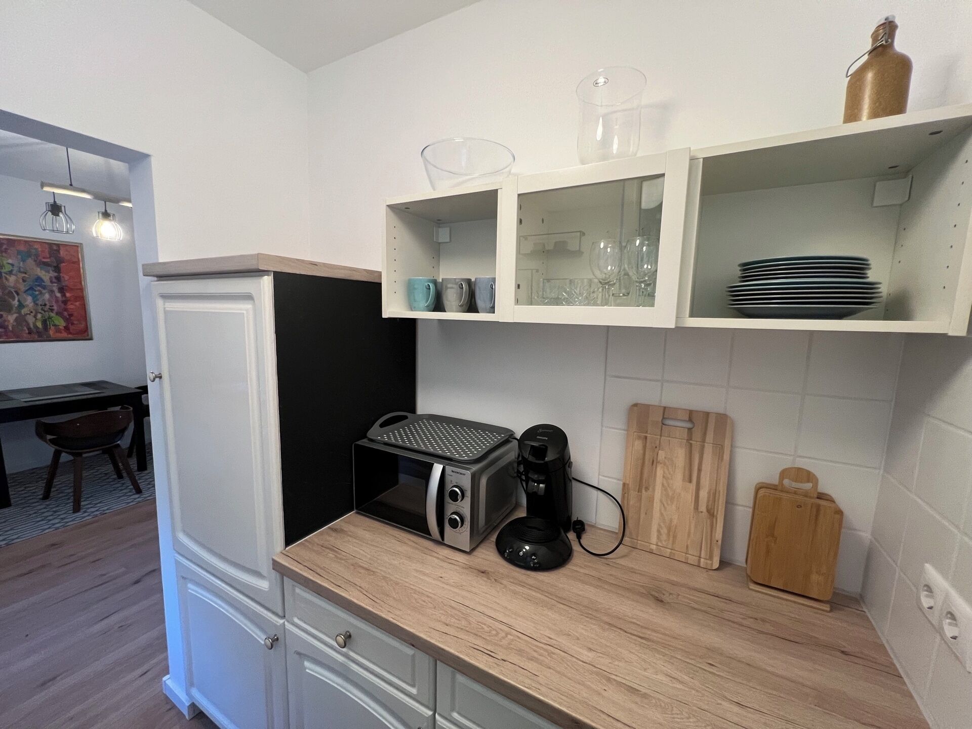 A compact kitchen with white cabinets, a built-in microwave, a coffee maker, and a wooden cutting board.