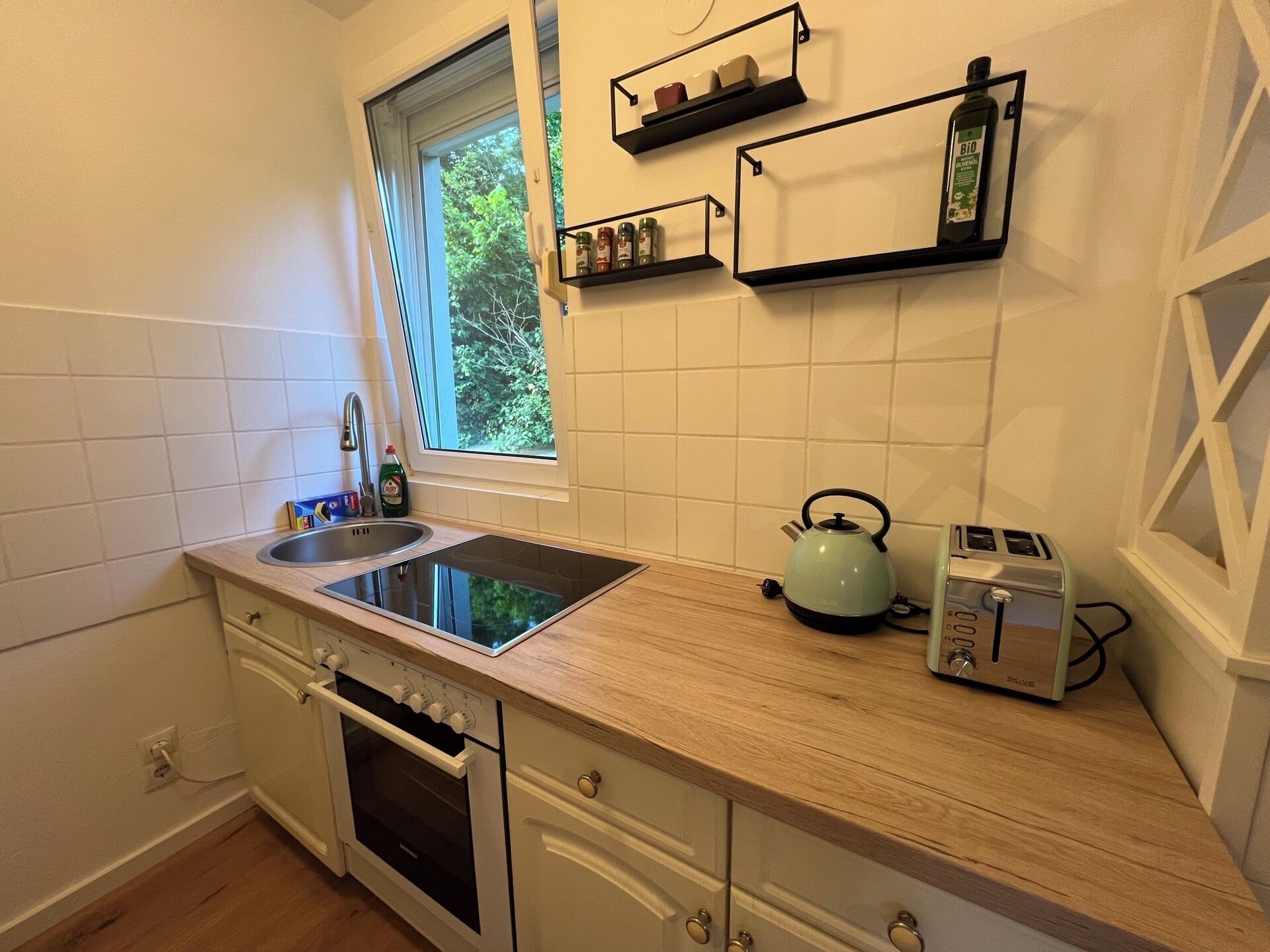 Una cocina con estufa integrada, un lavamanos y una ventana. Hay estantes sobre la encimera y una tetera en la encimera.