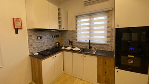 A modern kitchen with white cabinets, a black stove, and a window with blinds. - PON Bliss Apartments (Abuja)