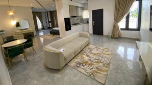 A modern living room with a sofa, dining table, and chairs. There is a kitchen area with cabinets and a stove. - PON Bliss Apartments (Abuja)