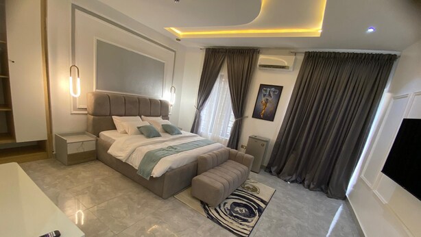 A modern bedroom with a large bed, bedside tables, a TV, and a remote control on a dresser. - PON Bliss Apartments (Abuja)