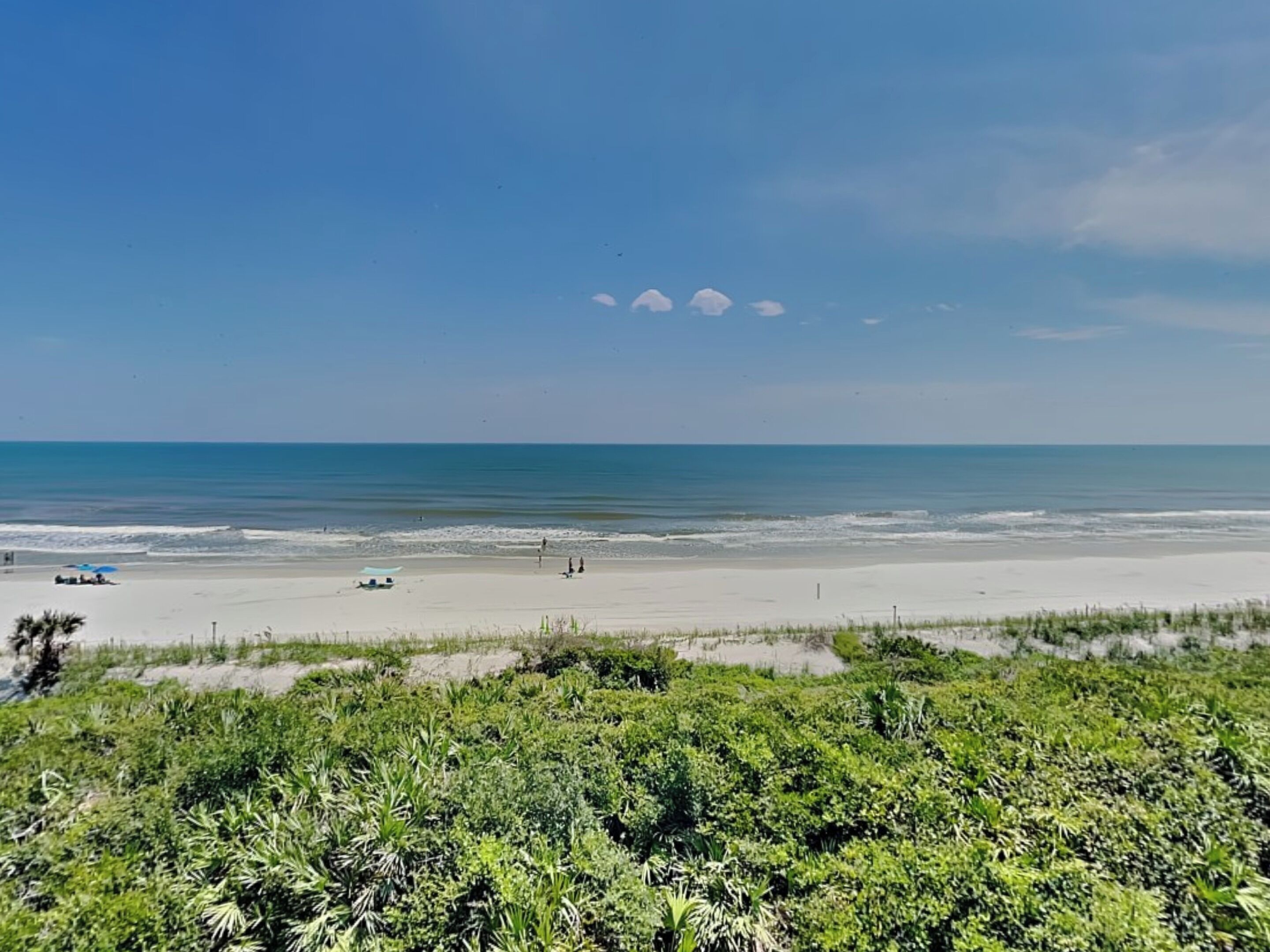 A beach with clear skies, calm waters, and sparse vegetation in the foreground.
