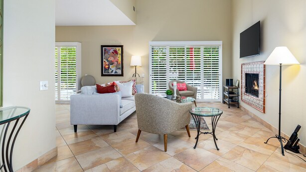 A spacious living room with a sofa, armchair, and glass coffee table, a fireplace, and a TV mounted on the wall.