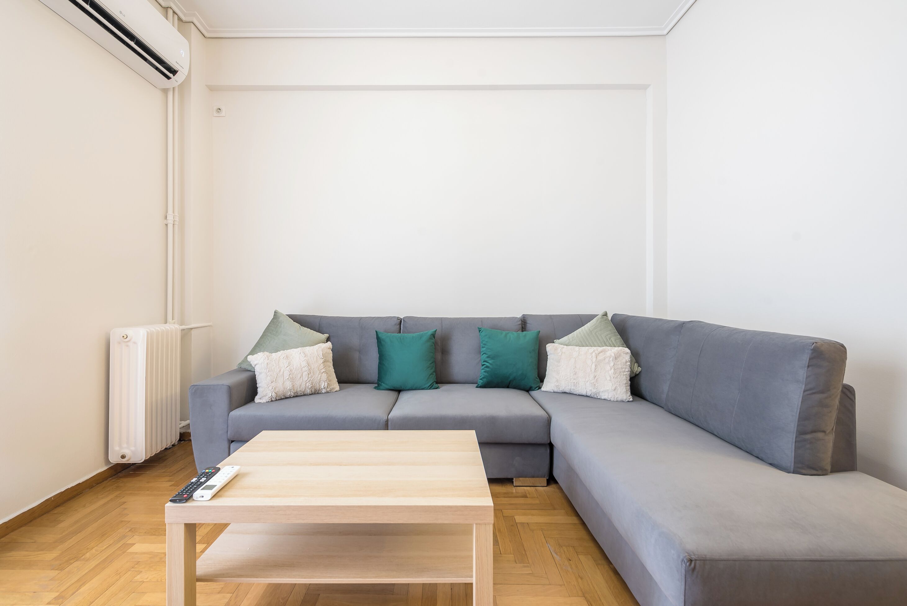 Un salon avec un canapé gris, deux coussins verts, une table basse en bois et un radiateur blanc.
