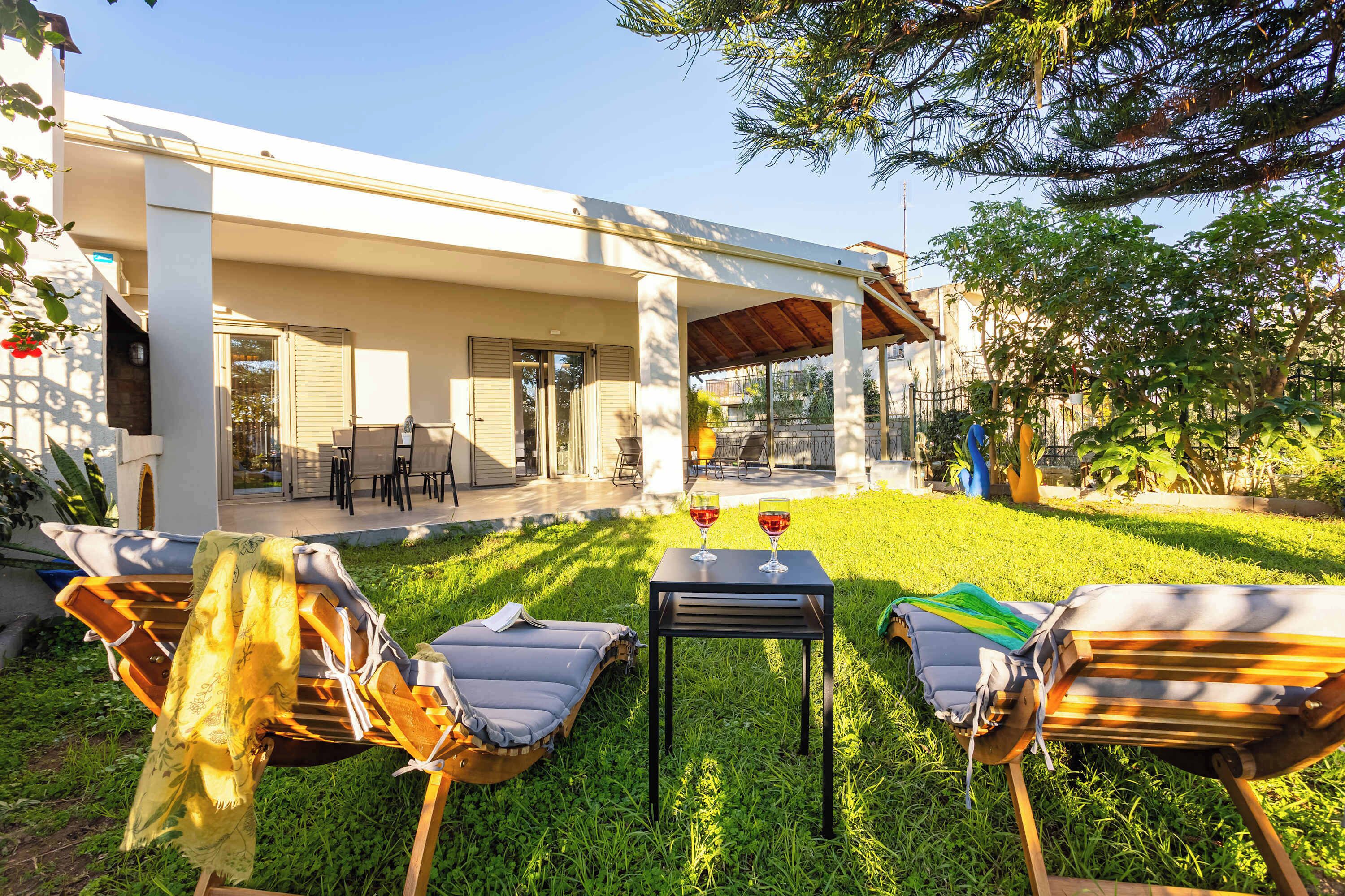 Et overdækket opholdsområde med loungesæder og et bord, en terrasse med udsigt til huset, og en velplejet græsplæne.