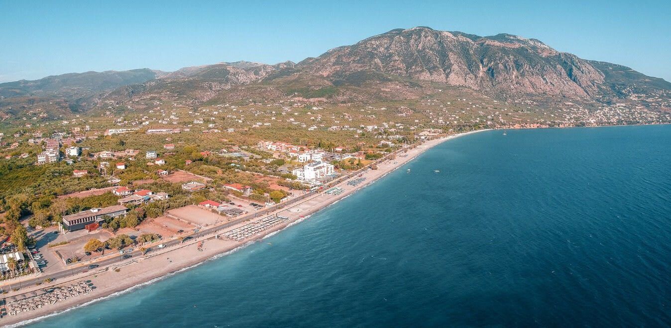 Vista aèria d'una població costanera amb una muntanya al fons, una platja i una carretera que segueix la línia de la costa.