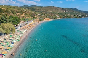 A coastal beach with clear turquoise waters, a row of beach umbrellas, and a hilly landscape in the background.