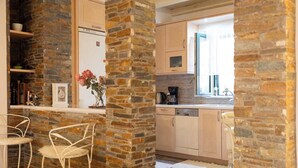 A kitchen with stone walls, wooden cabinets, and a window. There is a bar area with chairs and a vase of flowers. - Seafront Verga Stone Haven - Waves' Embrace (Paralia Vergas)