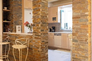 A kitchen with stone walls, wooden cabinets, and a window. There is a bar area with chairs and a vase of flowers.