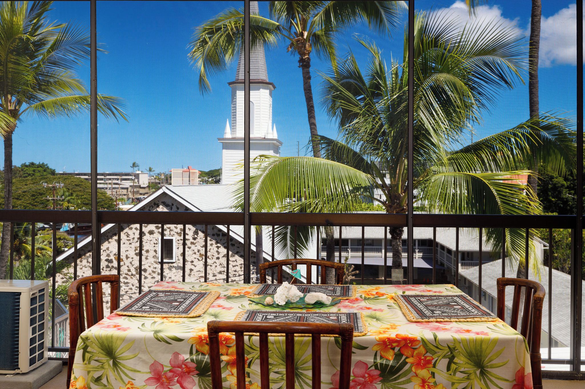 A table with a floral tablecloth, two chairs, and a decorative centerpiece on a balcony with a view of a white building and palm trees.