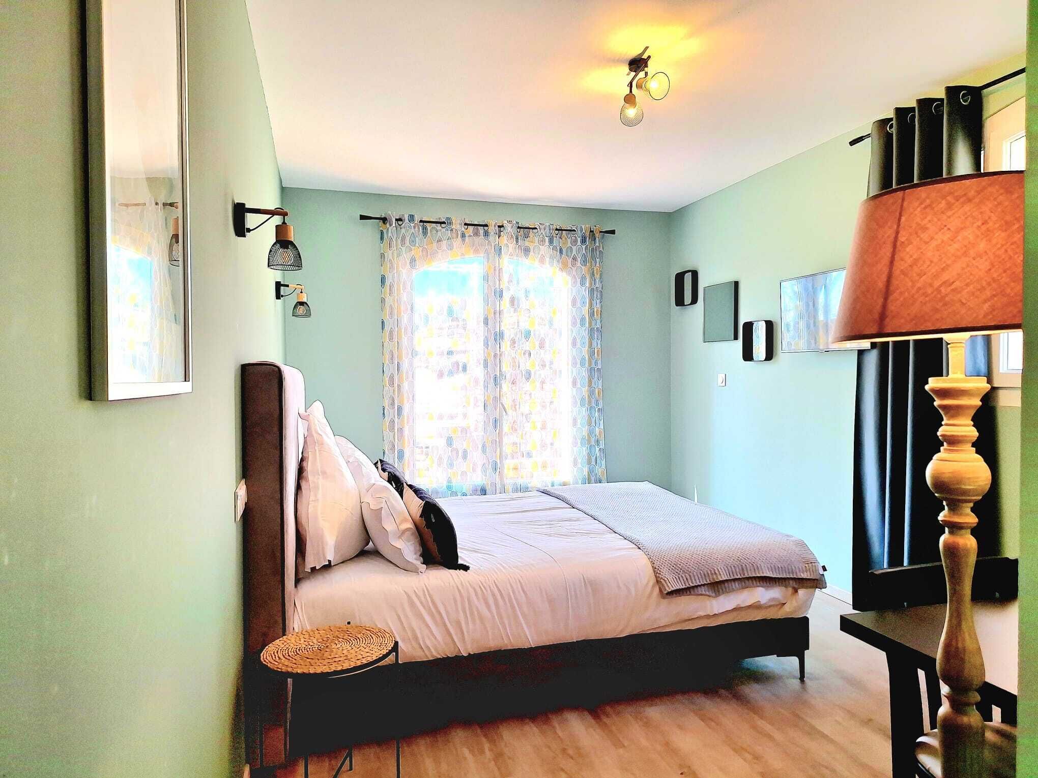 A bedroom with a bed, a bedside lamp, a wall-mounted TV, and a window with curtains.