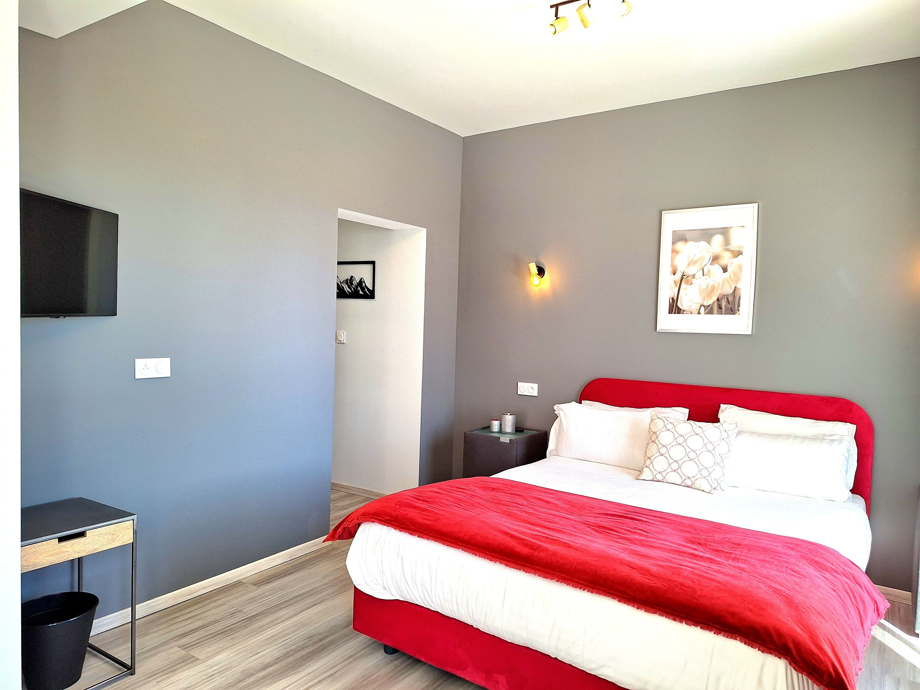 A bedroom with a red bed, grey walls, a mounted TV, and a framed picture on the wall.