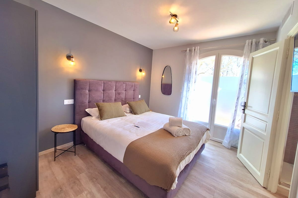 A bedroom with a large bed, a small side table, and a view of the outdoors through a door.