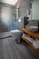 Un baño moderno con ducha de vidrio, inodoro blanco, tocador de madera con lavamanos grande y toallero.