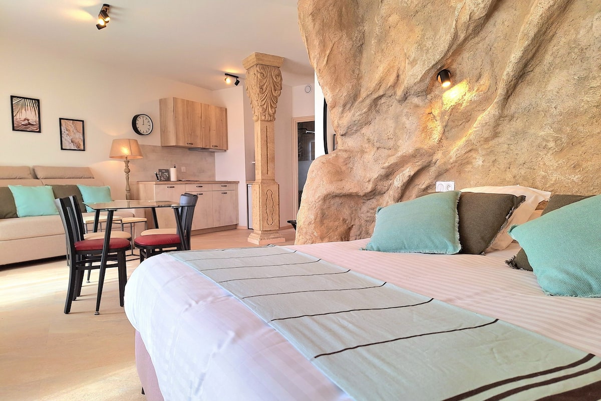 A bedroom with a large bed, a dining area with a table and chairs, a sofa, and a kitchen in the background.