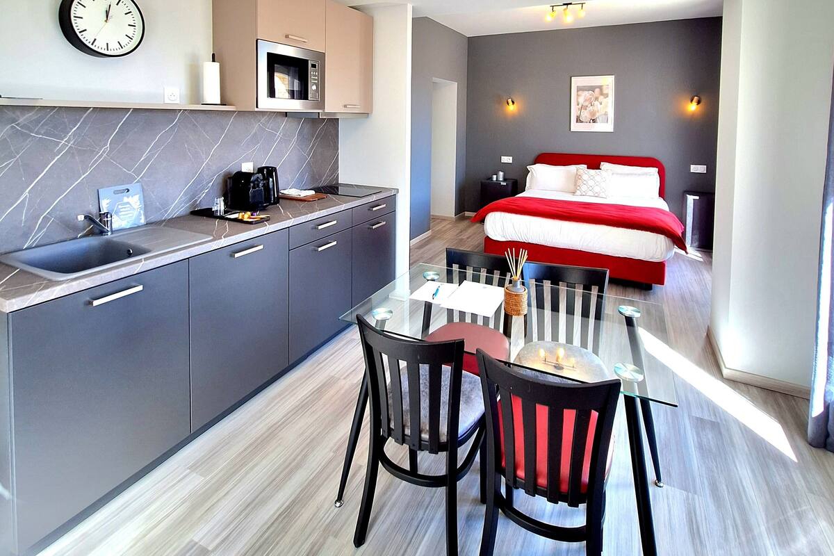 A modern kitchen with a built-in microwave, a dining area with a glass table and chairs, and a bedroom with a red bed and white pillows.