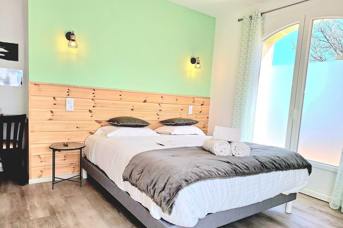 A neatly made bed with a wooden headboard, a small bedside table, and a view of the outdoors through a window with curtains.