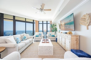 A spacious living room with a large window, a ceiling fan, a flat-screen TV, and a view of the ocean. - Phoenix West 2704 3 Bedroom Condo by RedAwning (Orange Beach)