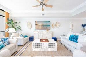 A living room with a ceiling fan, a large flat-screen TV, and a decorative wall with leaf patterns. - Phoenix West 2704 3 Bedroom Condo by RedAwning (Orange Beach)