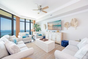 A spacious living room with a large window, a ceiling fan, a white sofa set, a coffee table, and a wooden cabinet. - Phoenix West 2704 3 Bedroom Condo by RedAwning (Orange Beach)