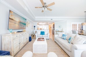 A spacious living room with a large TV, a ceiling fan, and a kitchen in the background. - Phoenix West 2704 3 Bedroom Condo by RedAwning (Orange Beach)