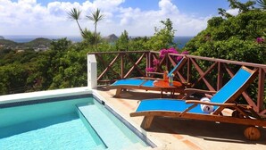 A poolside area with two lounge chairs and a table, surrounded by lush greenery and a view of the ocean.