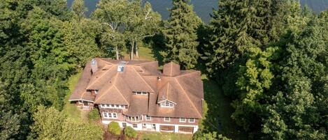 A large, multi-story house with a symmetrical roof and multiple chimneys, surrounded by dense forest and a body of water.