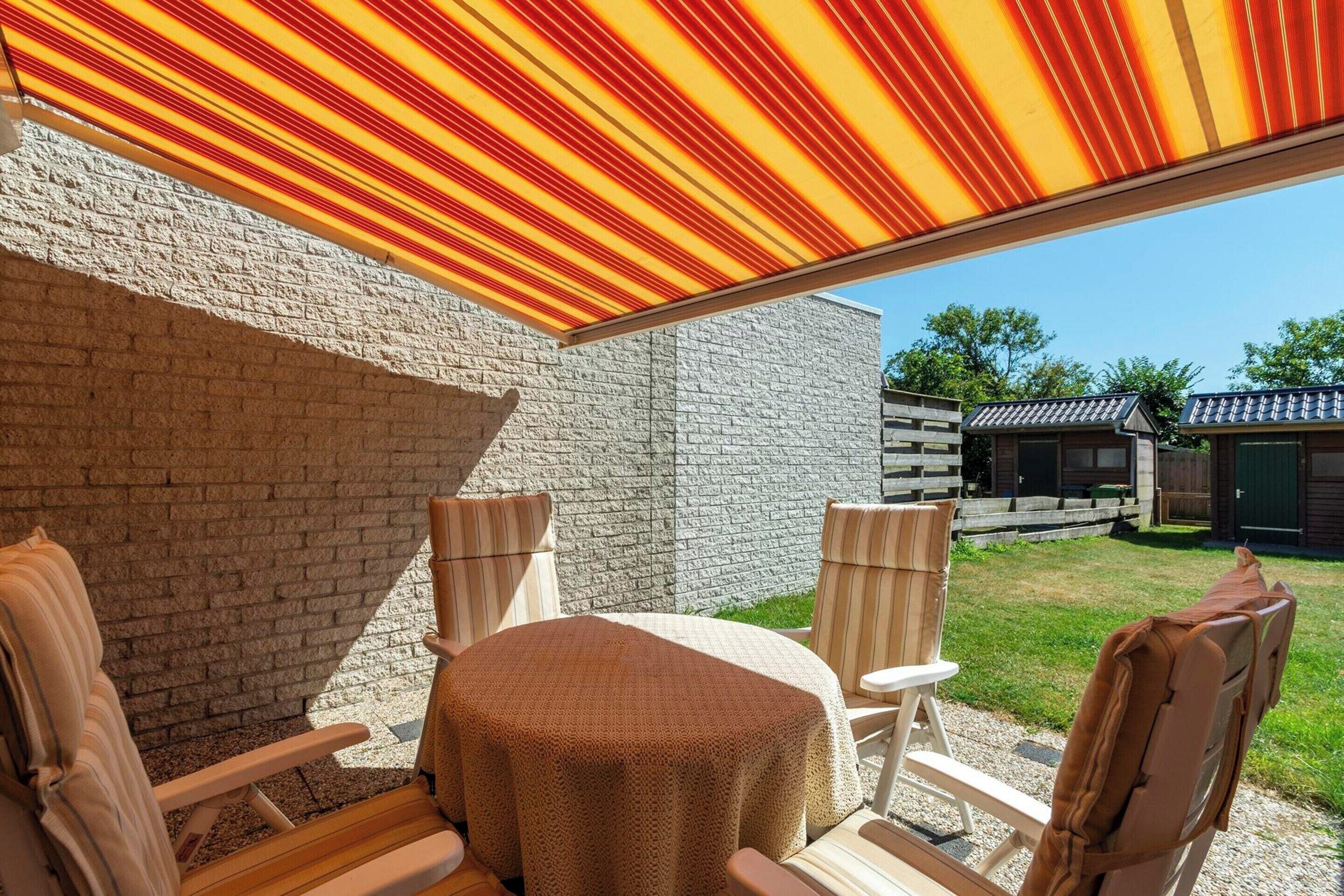 Patio esterno con tavolo e sedie, tenda a righe e parete in mattoni.