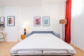 Ein Hotelzimmer mit Bett, Nachttisch mit Lampe, Schreibtisch mit Computer und zwei gerahmten Bildern an der Wand.