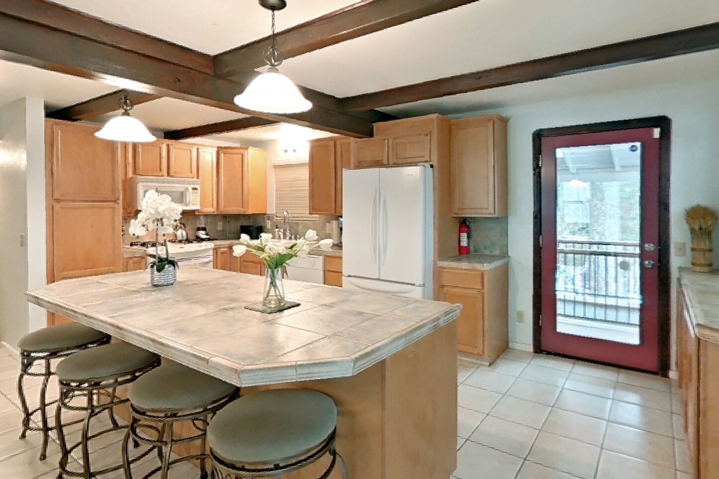 A kitchen with wooden cabinets, a central island with stools, a refrigerator, and a red door.