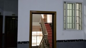 Ένα δωμάτιο με ξύλινη πόρτα, ένα παράθυρο με κάθετες μπάρες και μια σκάλα που οδηγεί σε έν αν άλλο χώρο.