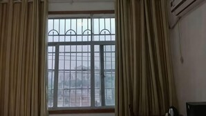 Ένα δωμάτιο με ένα μεγάλο παράθυρο, κουρτίνες, μια μονάδα κλιματισμού και μια καρέκλα.