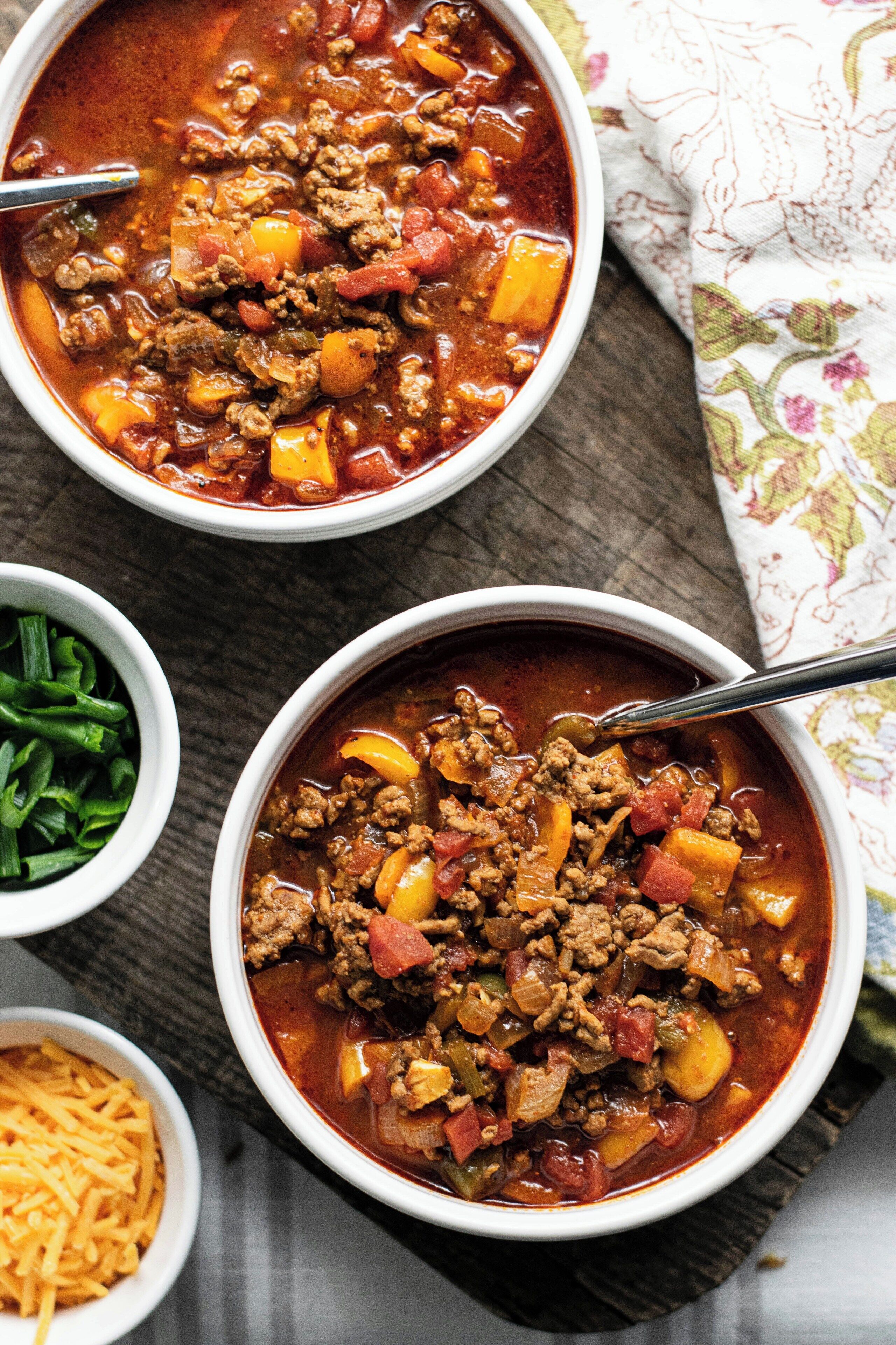 Two bowls of chili with visible ingredients like ground meat, tomatoes, and peppers, served on a wooden table with a cloth napkin.