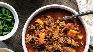 Two bowls of chili with visible ingredients like ground meat, tomatoes, and peppers, served on a wooden table with a cloth napkin.