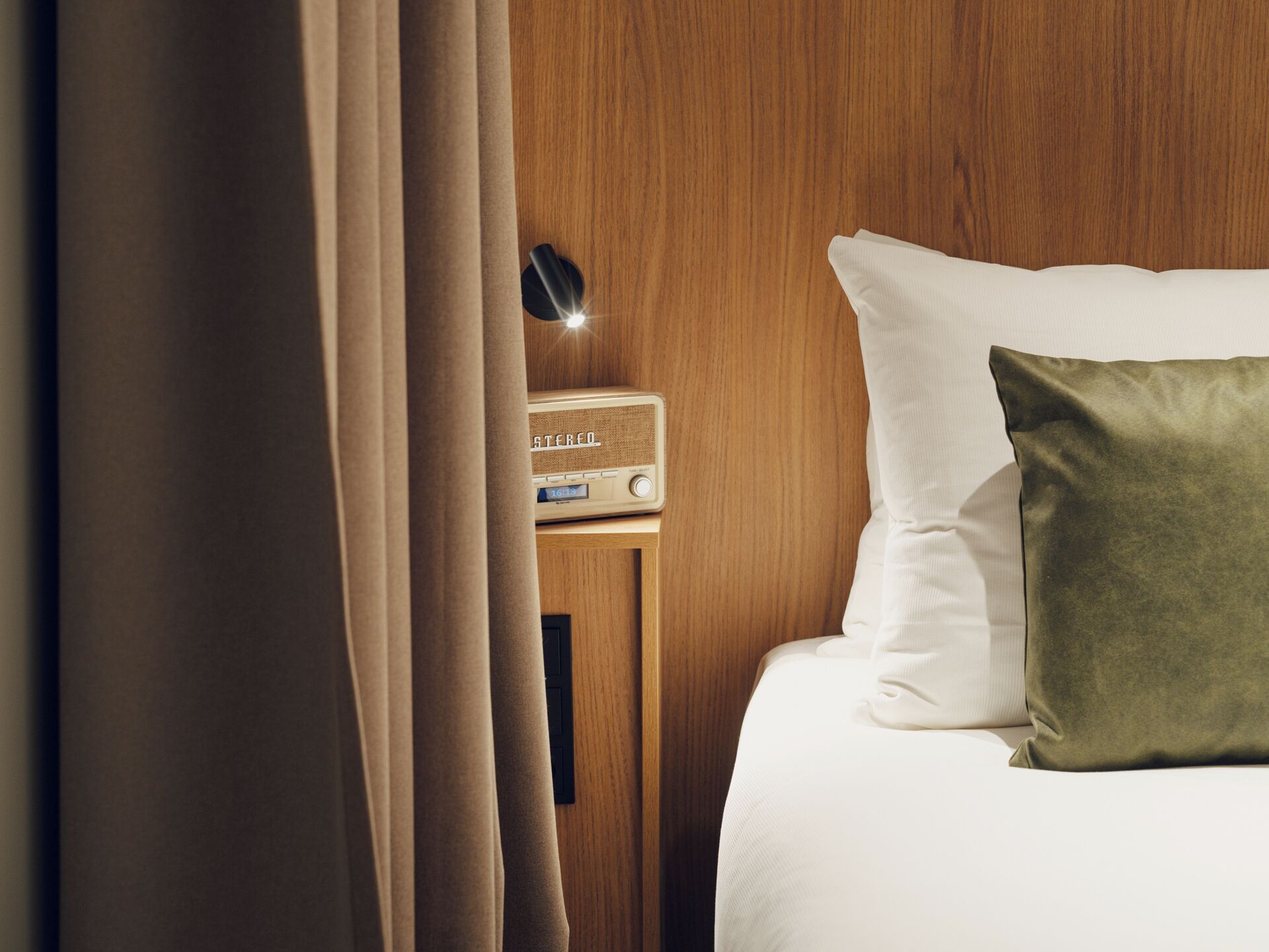 Una cama bien hecha con almohadas blancas y verdes, una mesita de noche de madera con una radio y una cortina entreabierta.