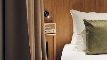 Una cama bien hecha con almohadas blancas y verdes, una mesita de noche de madera con una radio y una cortina entreabierta.