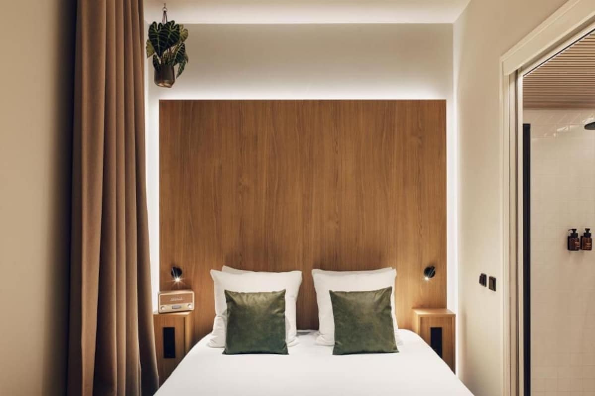 A hotel room with a wooden headboard, two bedside tables with lamps, a bed with white linens and green pillows, a hanging plant, and a view of the bathroom.