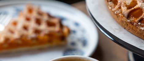 Una taza de café con arte de latte en el platito, un gofre en un plato, y una porción de pastel en el fondo.