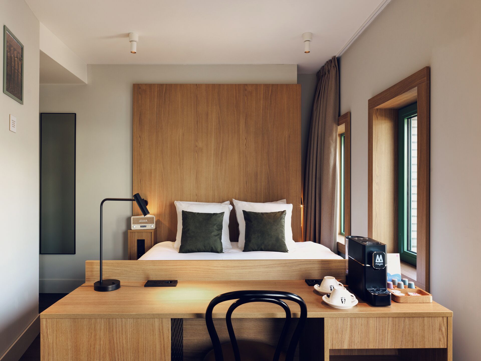 Habitación de hotel con cabecera de madera, cama con dos almohadas, escritorio con cafetera y una silla.