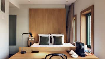 Habitación de hotel con cabecera de madera, cama con dos almohadas, escritorio con cafetera y una silla.