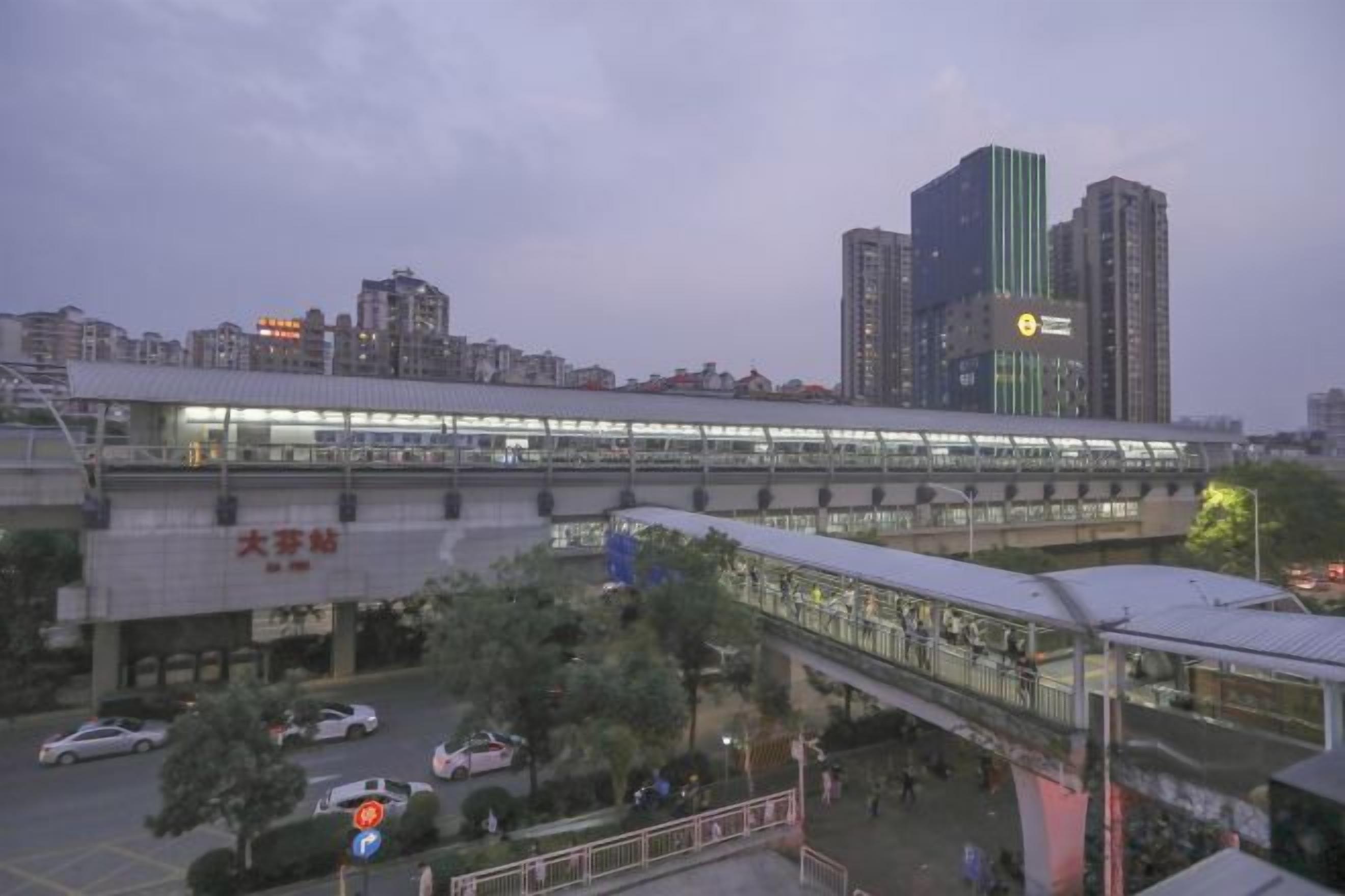 A train station with a pedestrian bridge, a parking area, and a cityscape in the background.