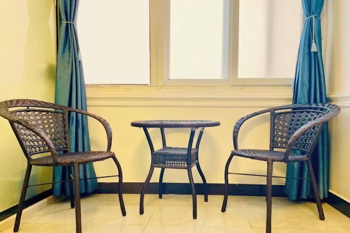 Una esquina con dos sillas de mimbre y una mesita redonda, una ventana con cortinas y una pared lisa.
