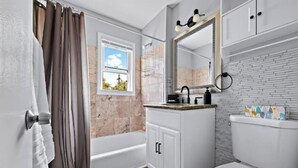 A bathroom with a bathtub, a toilet, a vanity with a mirror, and a window with a view of the sky.