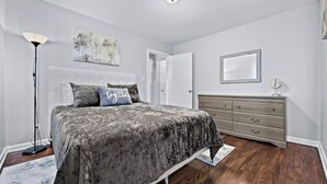 A bedroom with a bed, bedside lamp, dresser, and artwork on the wall.