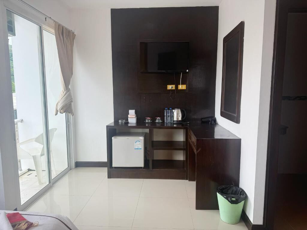 A compact hotel room with a kitchenette, a TV mounted on the wall, a sliding glass door leading to an outdoor area, and a small trash bin.