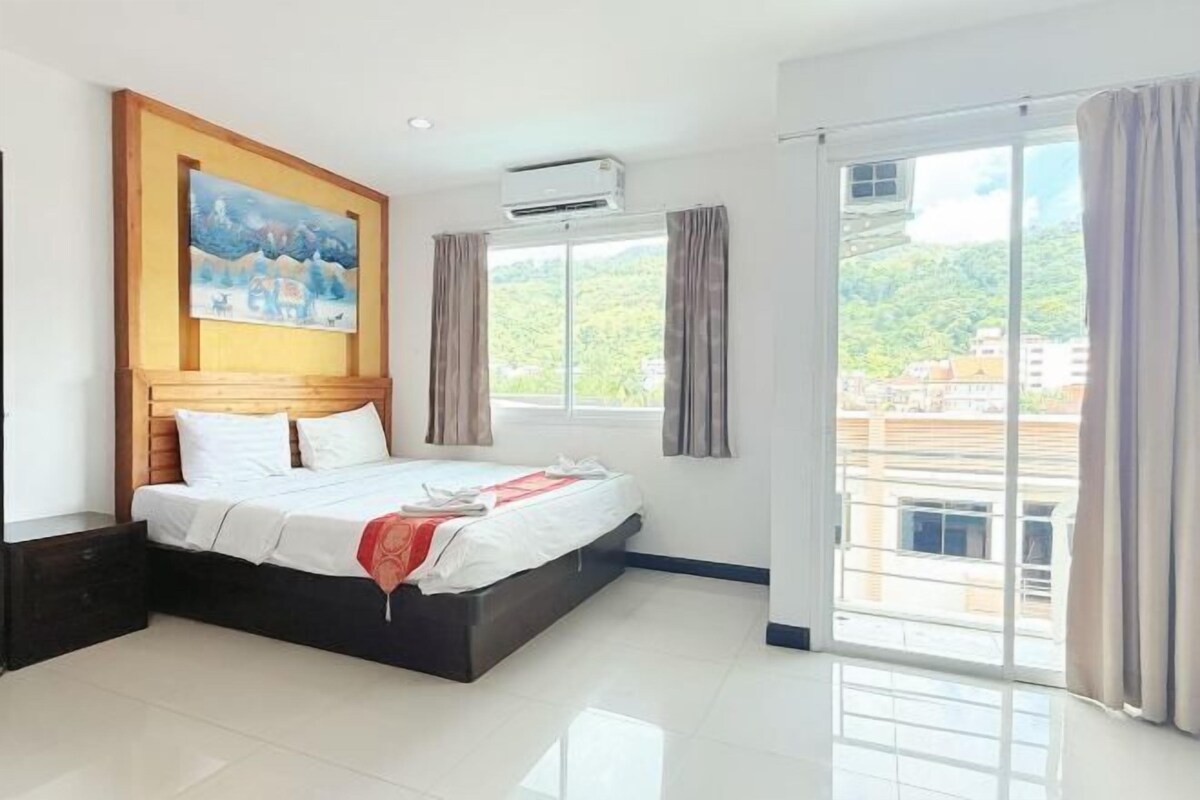 A hotel room with a bed, a painting, an air conditioner, a window with curtains, and a view of buildings.